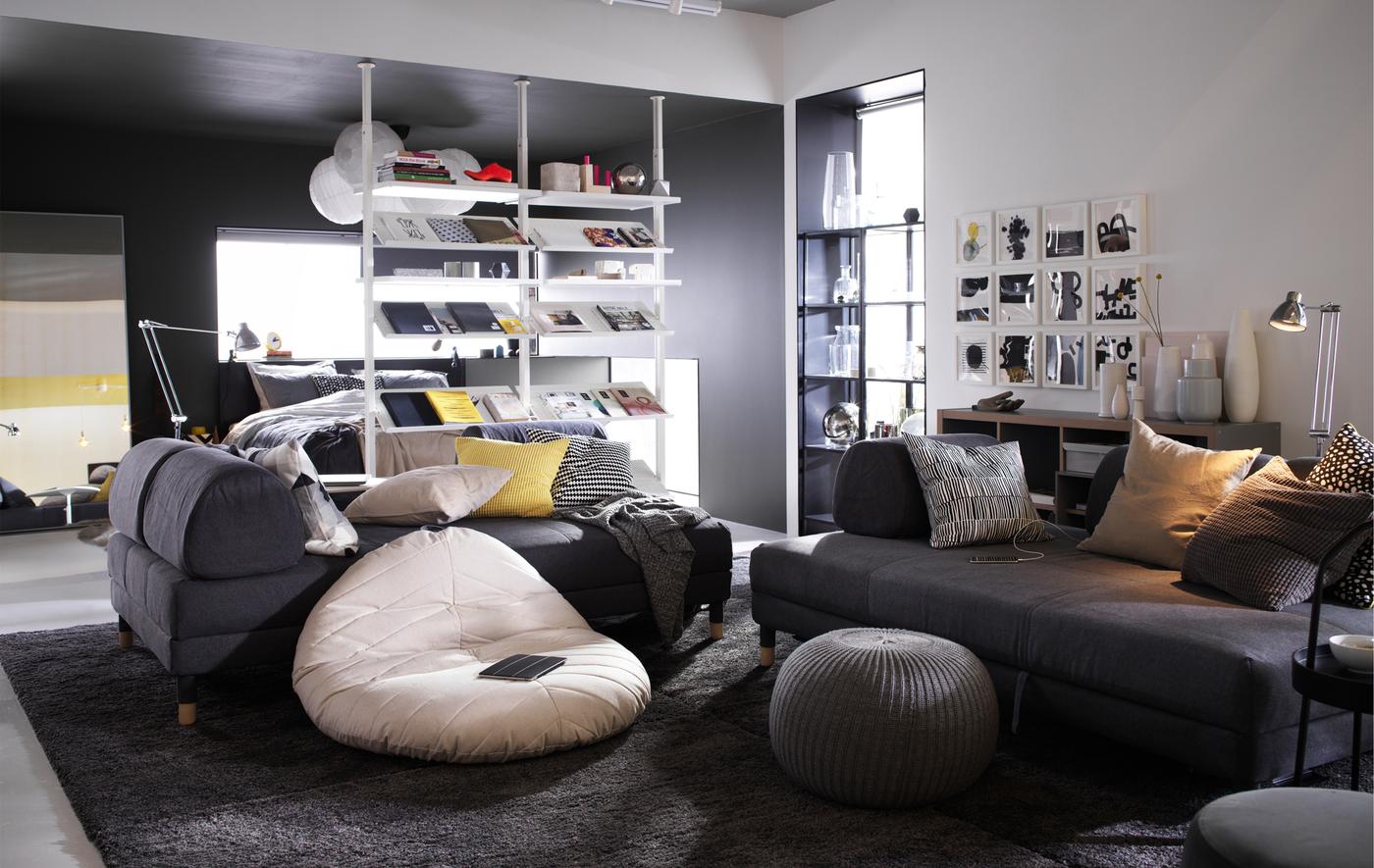 Un soggiorno sui toni del bianco e del grigio con un pouf DIHULT e due divani. Una combinazione ELVARLI lo separa dalla zona letto.