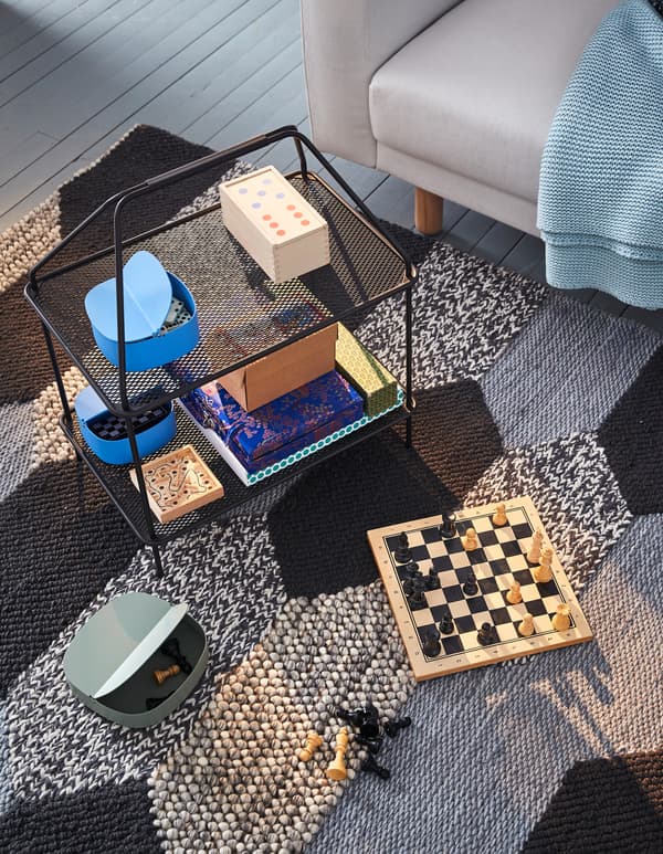 Un soporte de revistas IKEA YPPERLIG negro utilizado para colocar xogos de mesa sobre unha alfombra negra e gris.