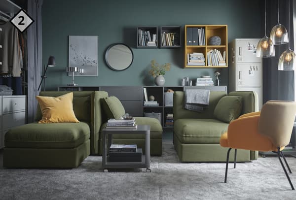 Un stanza con elementi angolari verdi, tavolino grigio e una poltrona bicolore. Dietro gli elementi angolari mobili scuri e lampade da tavolo. 