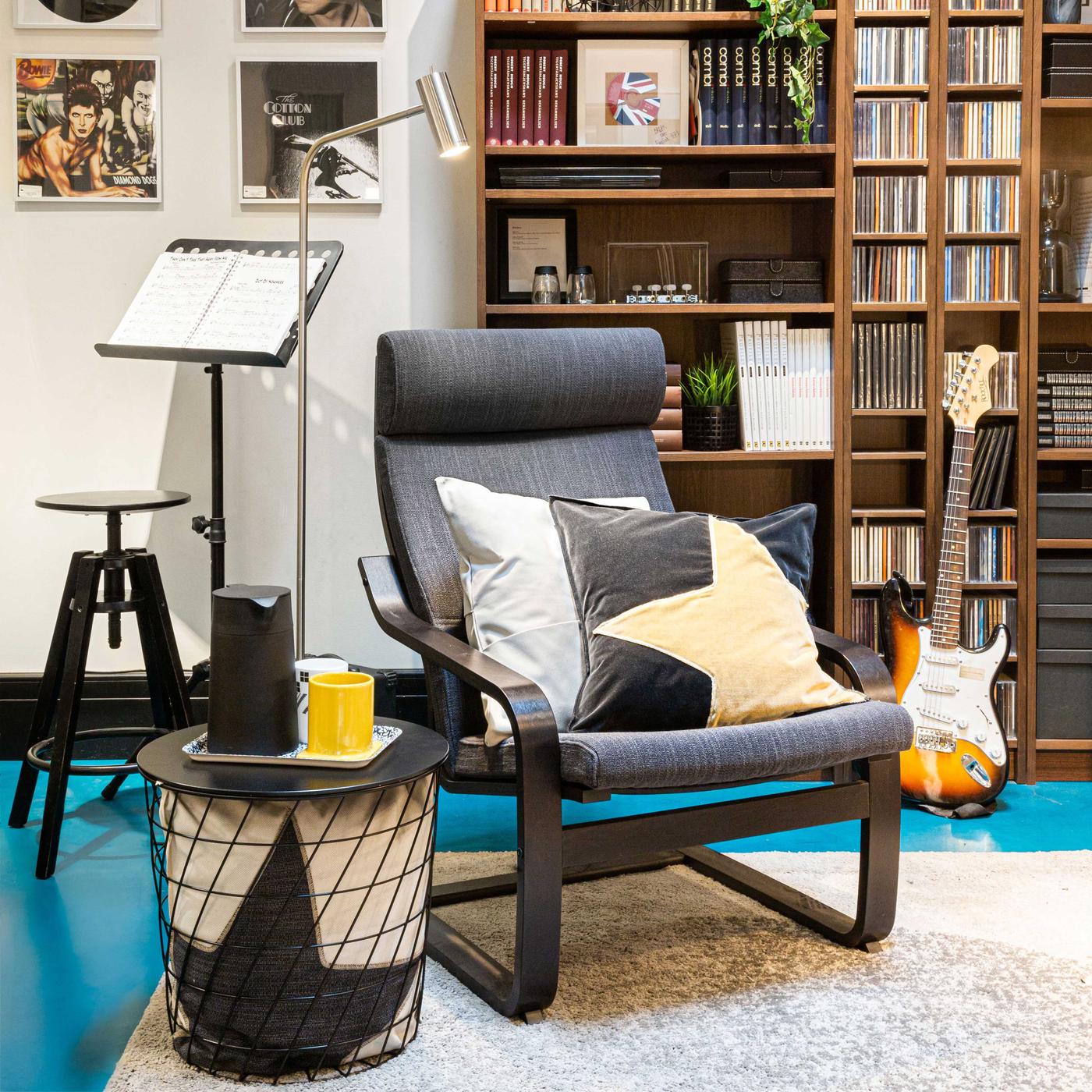Un stanza con poltrona POÄNG, un tavolino KVISTBRO, un libreria, una lampada da terra /lettura NYMÅNE, un chitarra elettrica e un tappeto. Sul tavolino è messo un vassoio con una brocca, un bicchiere e una tazza. A sinistra si vede uno sgabello, un leggio con uno spartito.