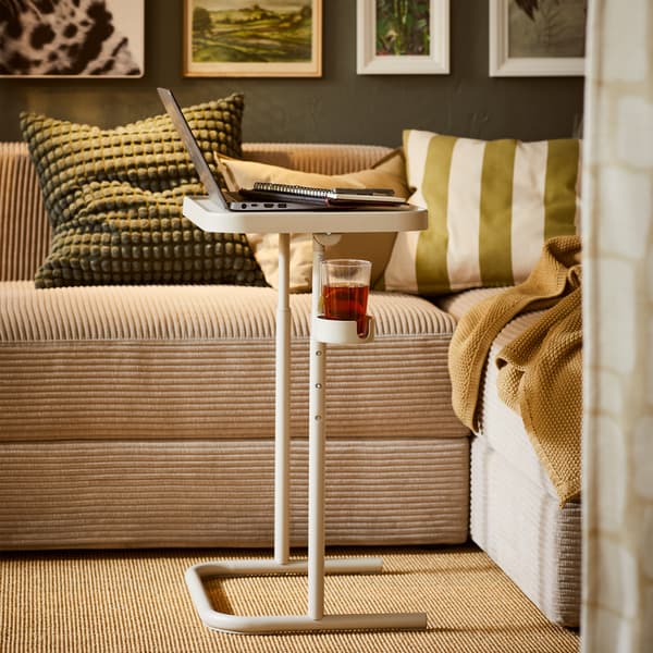 Un support pour ordinateur portable BJÖRKÅSEN beige situé près d’un canapé JÄTTEBO gris-beige agrémenté de textiles. Une rangée d’œuvres artistiques encadrées au-dessus du canapé.