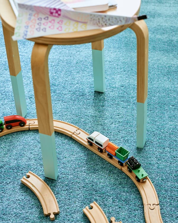 Un taburet din lemn cu partea de jos a picioarelor vopsită turcoaz este așezat pe un covor turcoaz. O șină de tren de jucărie șerpuiește printre picioarele sale.