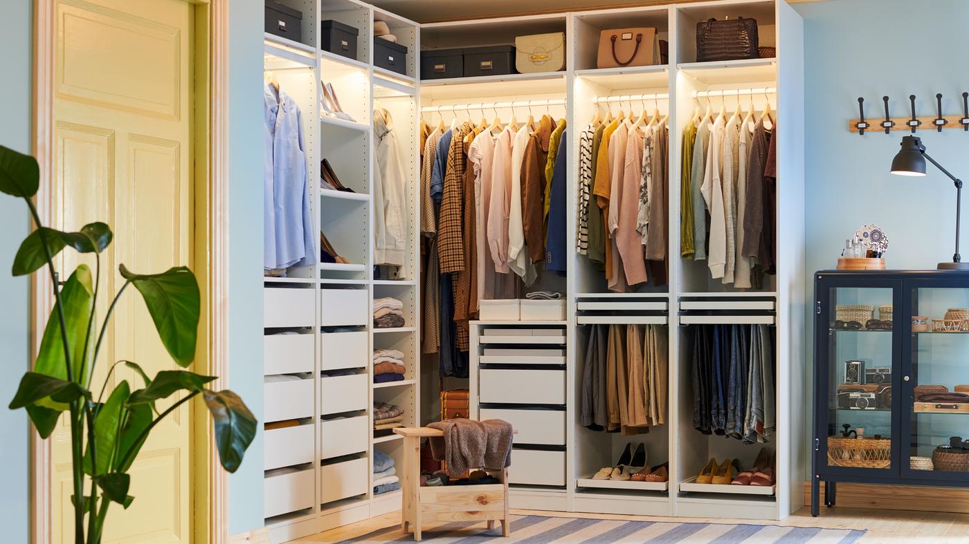 Un taburete con almacenaje PERJOHAN de pino delante de un gran armario de esquina PAX abierto con ropa, zapatos, cajas y bolsos.
