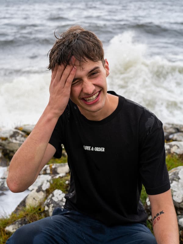 Un tânăr îmbrăcat cu un tricou negru stă la malul oceanului, pe o piatră, râzând. Valurile se sparg în spatele lui.