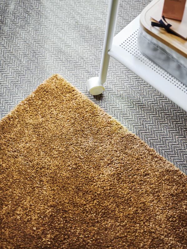 Un tapis STOENSE brun-jaune sur un sol gris, une desserte à roulettes en métal posée à côté.