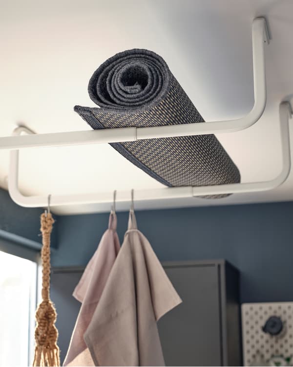 Un tappeto arrotolato MORUM appoggiato sopra dei bastoni appendiabiti fissati al soffitto di una veranda, degli strofinacci appesi sotto.