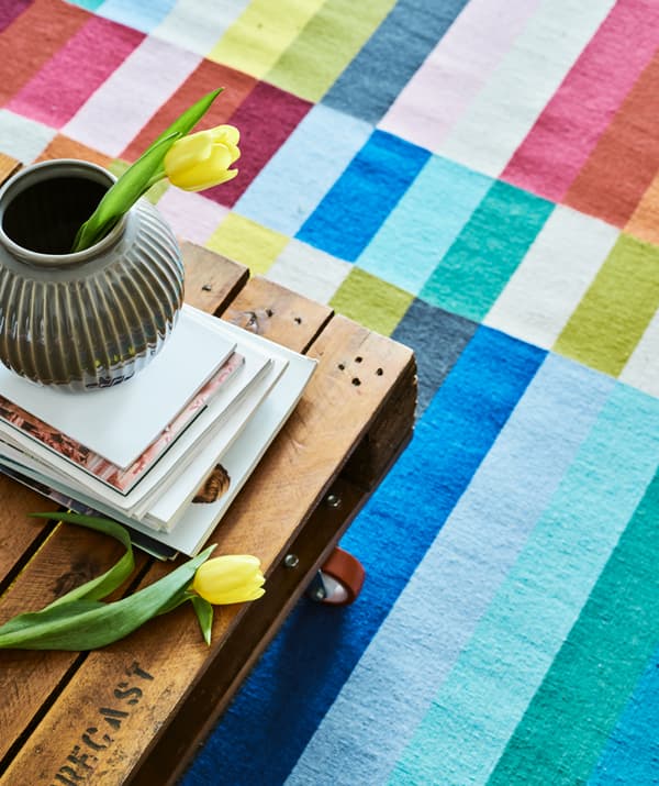 Un tavolino creato con un pallet riciclato e rotelle rosse su un tappetto a righe colorato. Sopra, riviste e un vaso grigio.