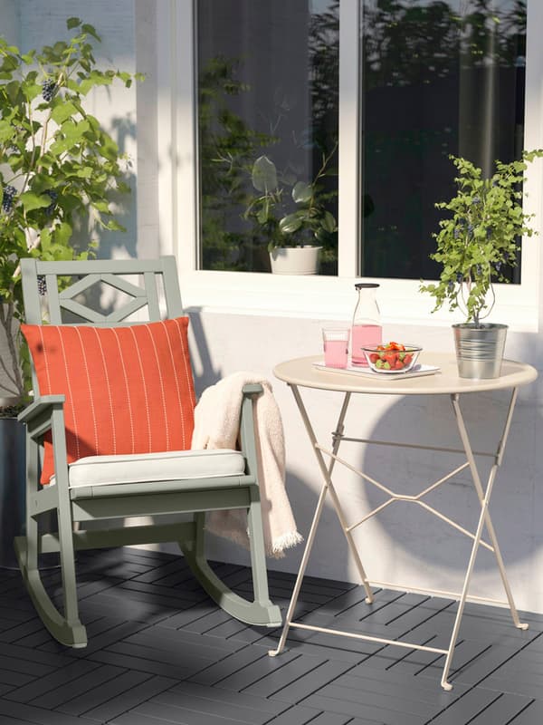 Un tavolino da esterno SUNDSÖ di colore bianco su cui poggiano una pianta, un bicchiere pieno, una bevanda in bottiglia e un ciotola di fragole. Accanto, c'è una sedia a dondolo.