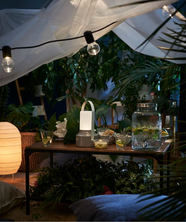 Un tavolino FJÄLLBO con sopra diverse decorazioni, bicchieri e un dispenser con rubinetto FÖRFRISKNING all'esterno copertoda da un telo e illuminato da diverse fonti di luce. 