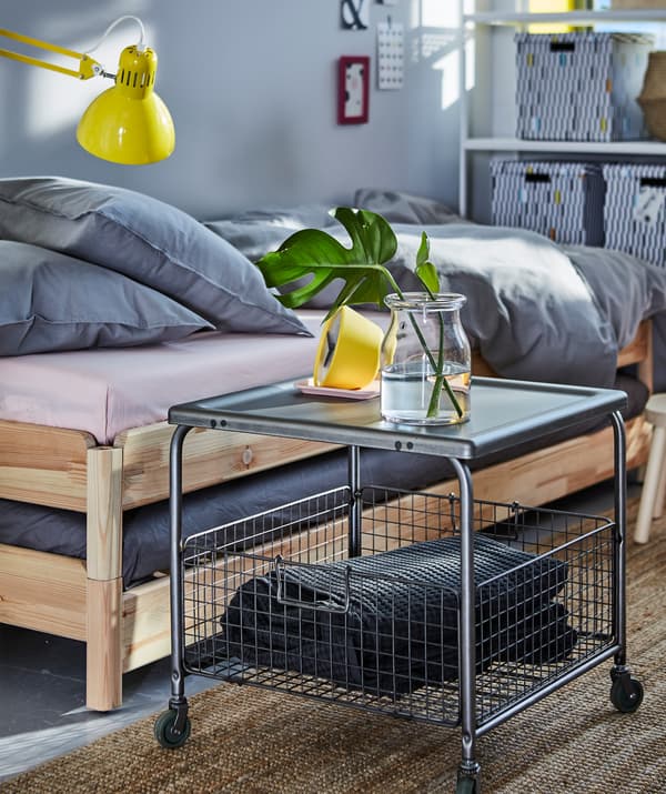 Un tavolino in acciaio grigio con contenitore in rete metallica con all'interno un plaid grigio. Sopra, un vaso in vetro con una pianta e una sveglia gialla.
