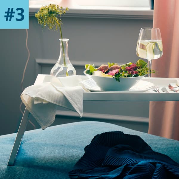 Un tavolino pieghevole da letto color bianco con una ciotola di insalata, un calice di vino e un vaso con fiori.
