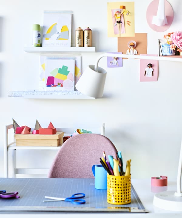 Un tavolo con un portapenne, una lampada da lavoro bianca, una sedia rosa e una parete con mensole per quadri dedicata alle creazioni artistiche - IKEA