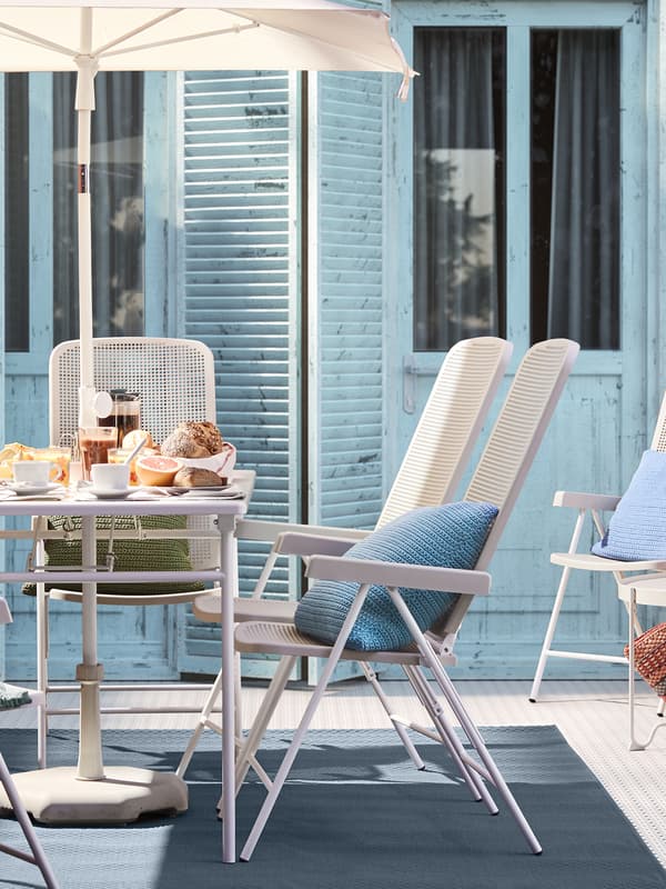 Un tavolo da esterno apparecchiato per la colazione con sedie e un ombrellone al centro. Sulle sedie ci sono cuscini colorati e sullo sfondo una portafinestra con ante azzure.