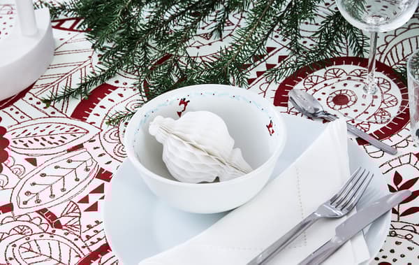 Un tavolo da pranzo apparecchiato per un’occasione di festa con tovaglia fantasia, candela a LED, ramoscello di abete e decorazioni in carta - IKEA