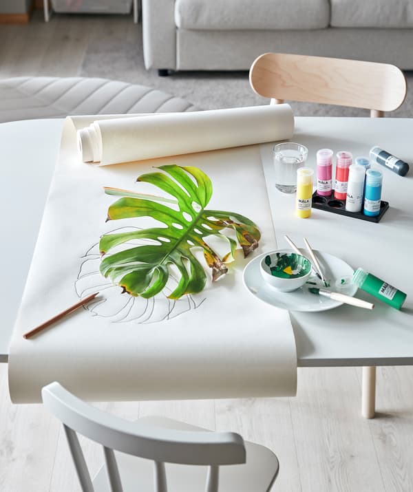 Un tavolo del soggiorno pronto per una sessione di pittura con le piante: colori MÅLA, carta da disegno e una foglia di MONSTERA parzialmente appassita.