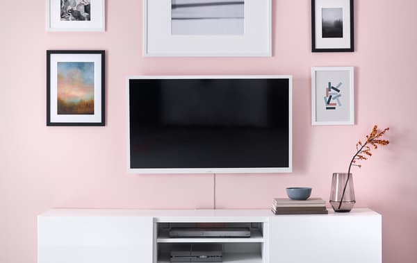 Un televisor muntat a una paret de sala rosa, al costat d'obres d'art a la paret i damunt un armari blanc.