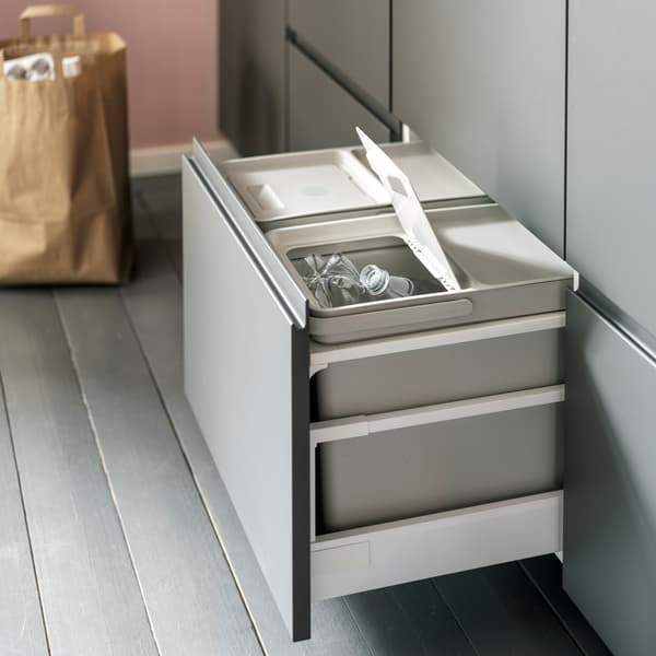 Un tiroir de cuisine ouvert avec des poubelles de tri des déchets gris clair à l’intérieur. Un couvercle est ouvert et des bouteilles en plastique sont triées à l’intérieur.