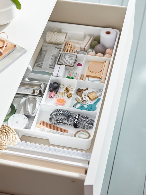 Un tiroir ouvert avec un insert KUGGIS blanc à huit compartiments remplis de clés, pinces, ficelle et fournitures de bureau.