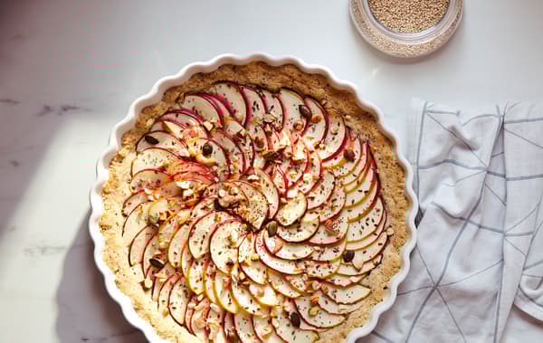 Un torchon de cuisine blanc orné d’un motif gris graphique et une tarte couverte de fines tranches de pommes dans un moule à tarte VARDAGEN blanc.