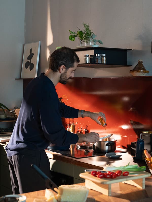 Un uomo ai fornelli versa un filo d'olio in una pentola, circondato da ingredienti freschi come pomodorini e formaggio.