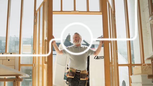 Un uomo con la barba è in piedi sulla soglia di una casa grezza in legno in costruzione, con entrambe le braccia appoggiate sugli stipiti della porta. Intorno alla sua testa è disegnato un cerchio dal quale partono due linee in direzioni opposte.