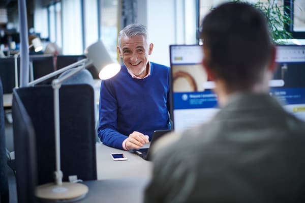 Un uomo con un pullover blu sta sorridendo dietro un computer e sorride a un'altra persona seduta di fronte a lui.