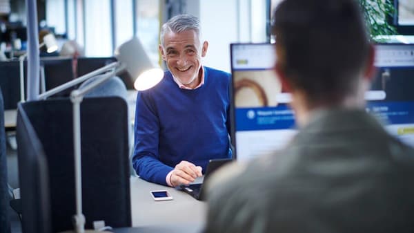 Un uomo con un pullover blu sta sorridendo dietro un computer e sorride a un'altra persona seduta di fronte a lui.