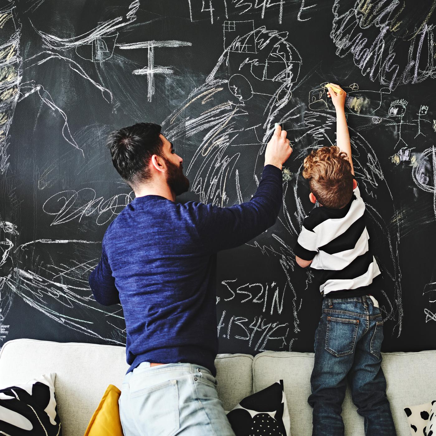 Un uomo e un bambino sopra un divano disegnano con il gesso su una parete dipinta con vernice effetto lavagna.