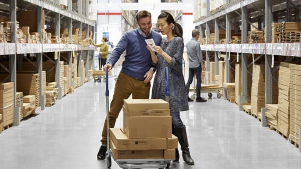 Un uomo e una donna guardano uno smartphone nell'area di ritiro dei singoli componenti degli articoli IKEA. Sullo sfondo un cliente guarda uno scaffale.