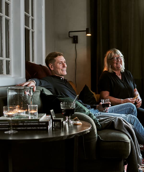 Un uomo e una donna su un divano verde con cuscini, plaid, bicchieri pieni, candeline e libri posati su un tavolino.