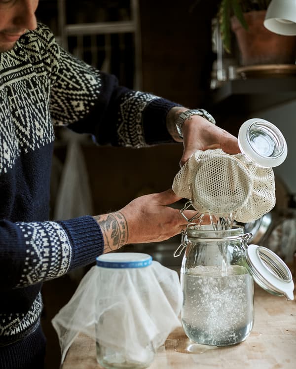 Un uomo lavora in cucina versando un liquido trasparente da un contenitore in stile Kilner a un altro utilizzando una tela di mussola come filtro - IKEA
