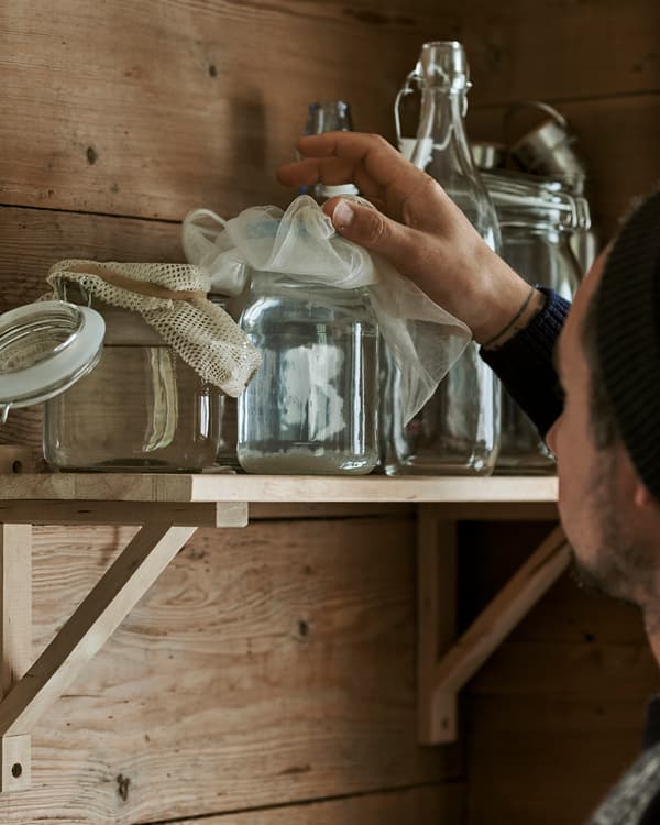 Un uomo prende da una mensola in legno un contenitore in vetro in stile Kilner, coperto da un piccolo scampolo di mussola - IKEA