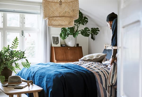Un uomo prepara il letto con biancheria a righe rosse, bianche e blu e un copriletto blu scuro in una stanza con una grande lampada a sospensione in bambù intrecciato - IKEA