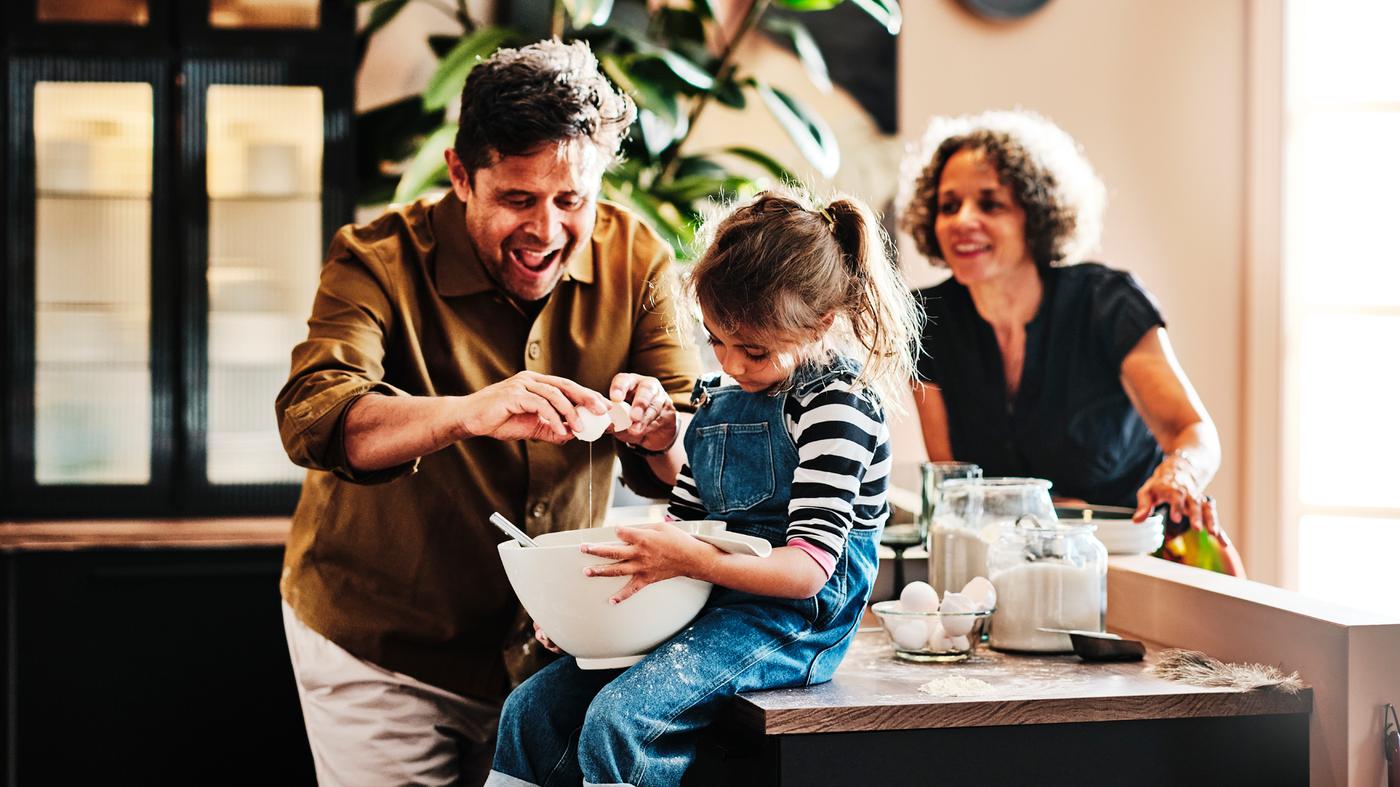 Un uomo rompe un uovo in una ciotola tenuta da una bambina che siede sul piano di lavoro della cucina, mentre una donna li guarda - IKEA.