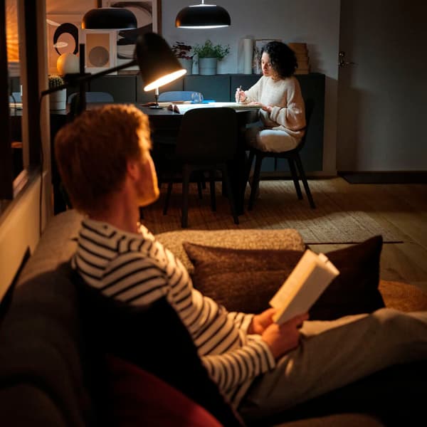 Un uomo seduto su un divano che tiene tra le mani un libro aperto e guarda una donna che lavora ad una scrivania sullo sfondo.