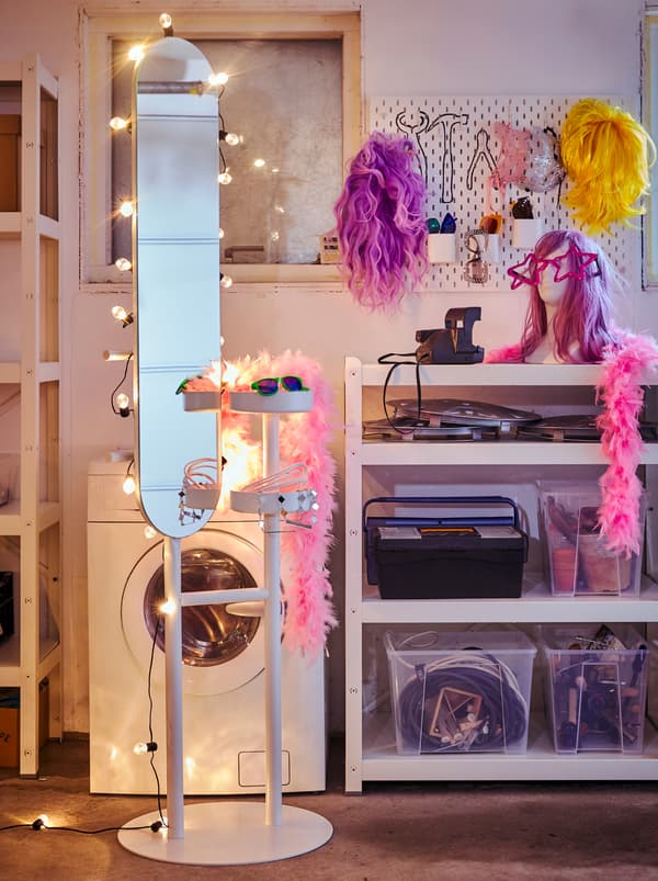 Un valet LIERSKOGEN décoré d’une guirlande lumineuse BLÖTSNÖ, se tenant près d’un panneau perforé SKÅDIS avec perruques et accessoires.