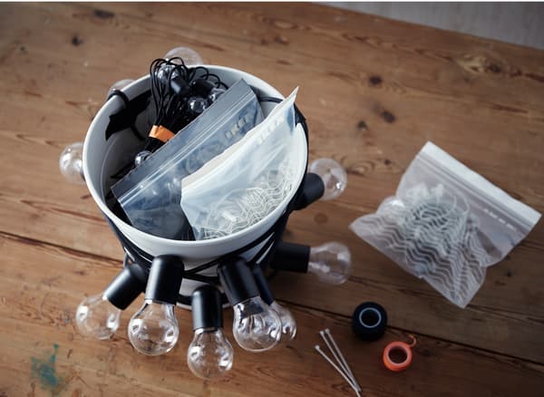 Un vaso bianco FNISS avvolto da cavi e lampadine e con al suo interno diverse buste e cavi.