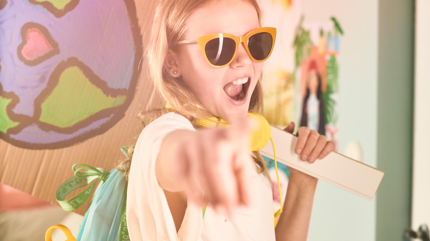 Una bambina, con occhiali da sole gialli, punta il dito mentre indossa uno zaino blu-verde BLÅVINGAD - IKEA.