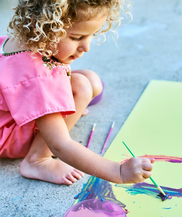 Una bambina dipinge con colori vivaci un foglio di carta gialla su un pavimento in cemento - IKEA