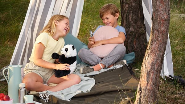 Una bambina e un bambino ridono insieme sotto una tenda realizzata con il tessuto a metraggio SOFIA a righe, seduti su una coperta per picnic FJÄLLMOTT - IKEA