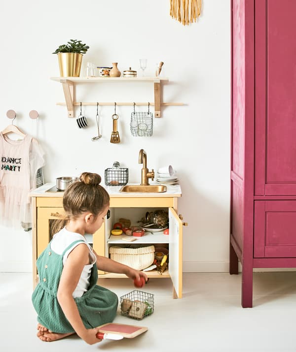 Una bambina gioca accanto a una cucina giocattolo con accessori come pentolini, cibo e piccoli utensili appesi a una mensola.