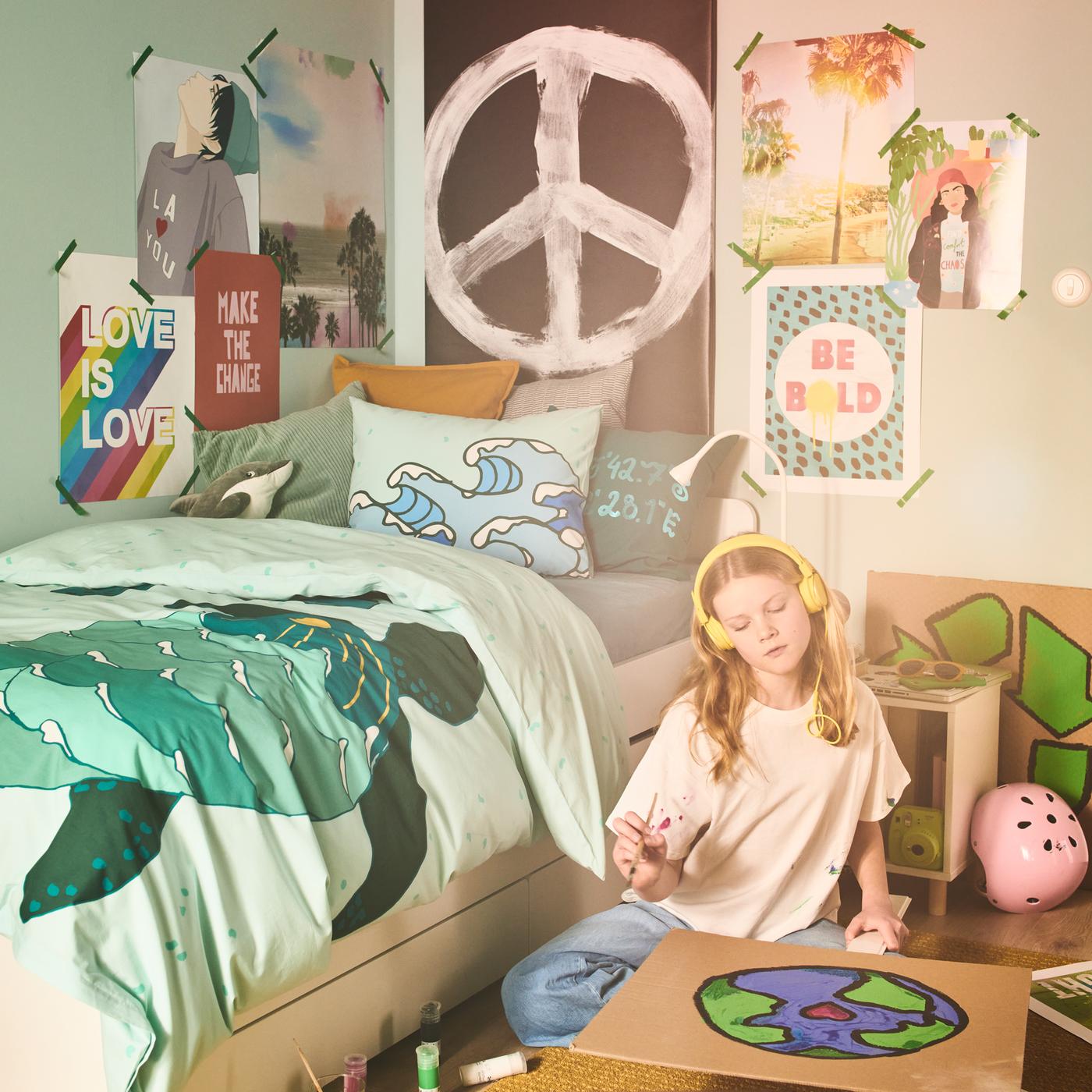 Una bambina intenta a colorare un poster del mondo è seduta sul pavimento accanto a un letto preparato con un copripiumino con federa e motivo tartaruga BLÅVINGAD.