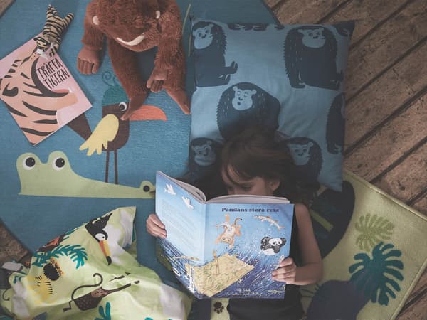 Una bambina mentre sta leggendo un libro sopra alcuni cuscini, tessili e peluche della collezione DJUNGELSKOG ispirata al tema della giungla.