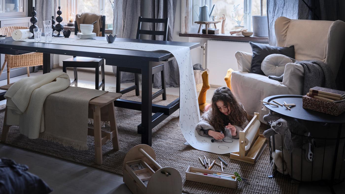 Una bambina, sdraiata su un tappeto, disegna su un rotolo di carta da disegno con un’estremità su un tavolo allungabile NORDVIKEN nero.