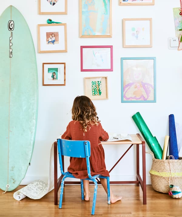 Una bambina seduta a una piccola scrivania su una sedia azzurra, sullo sfondo di una parete con disegni e foto di famiglia, accanto a una tavola da surf - IKEA