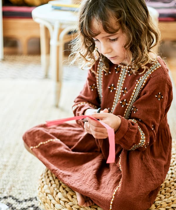 Una bambina seduta su un poggiapiedi in rattan mentre taglia un pezzo di carta - IKEA