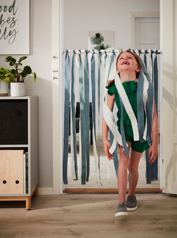 Una bambina sorridente passa sotto un bastone per tenda doccia BOTAREN decorato con strisce di tessuto DITTE appese alla soglia - IKEA