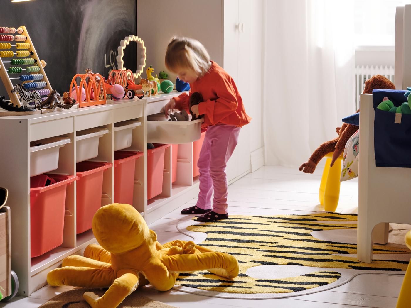 Una bambina, su un tappeto IDROTTSHALL giallo e nero, prende alcuni giocattoli da un contenitore in una combinazione di mobili TROFAST.