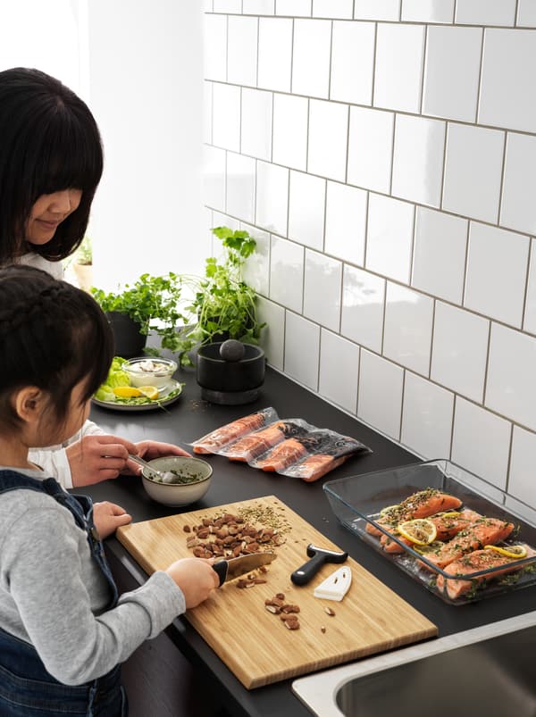 Una bambina usa un coltello SMÅBIT per affettare mandorle sul piano di lavoro nero di una cucina, aiutando la mamma a preparare una ricetta con il salmone.