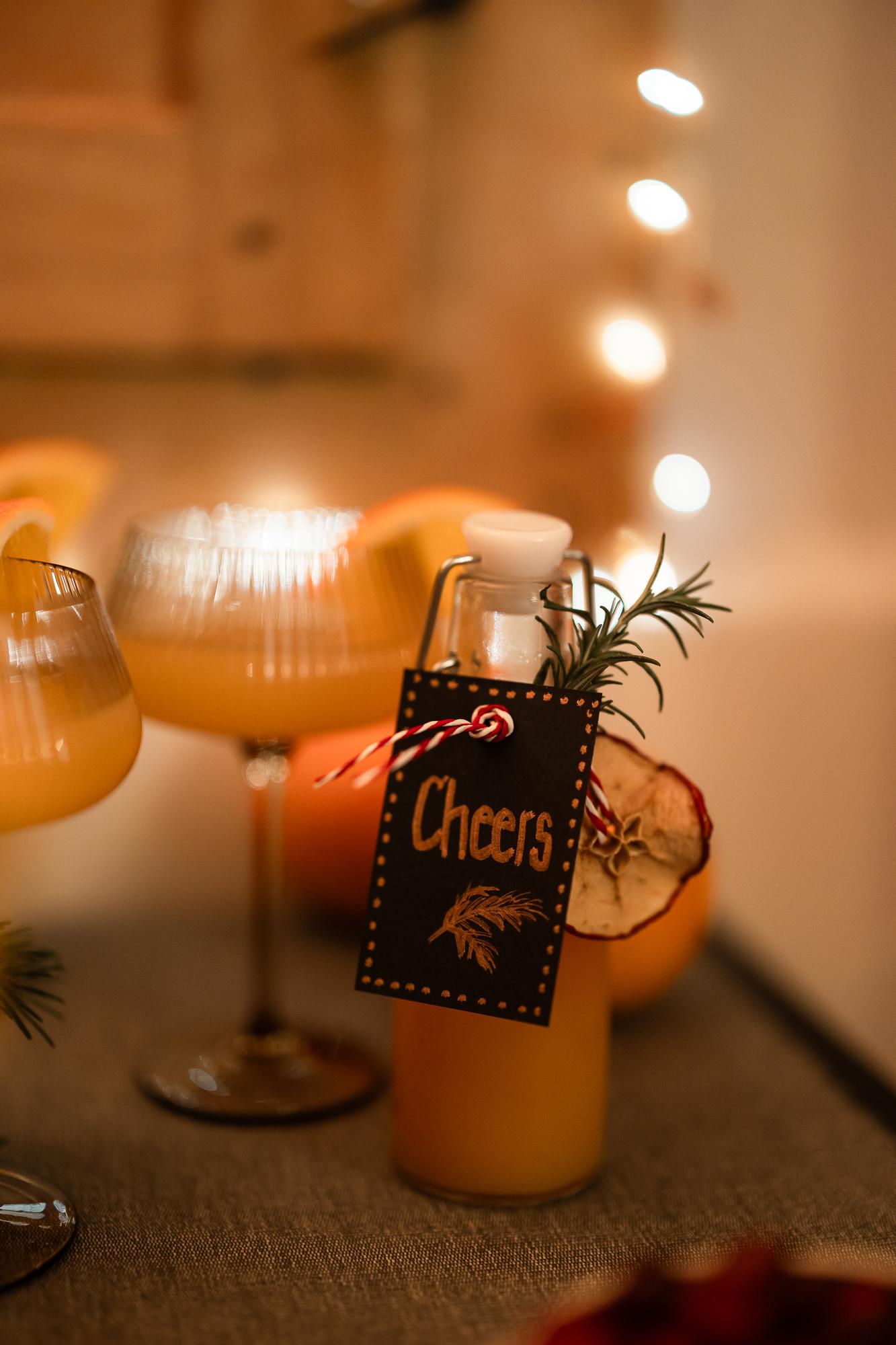 Una bottiglia di vetro con un'etichetta "Cheers" riposa tra bicchieri da cocktail, guarniti con frutta secca e rosmarino, sullo sfondo di luci festive.